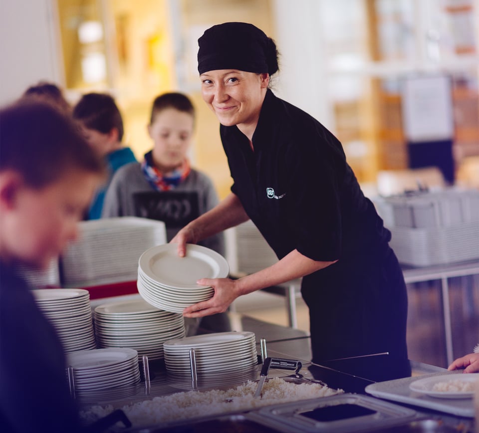 ISS:n ravintolatyöntekijä täydentämässä lautasia linjastoon koulun ruokasalissa, taustalla ruokaa noutavia koululaisia.