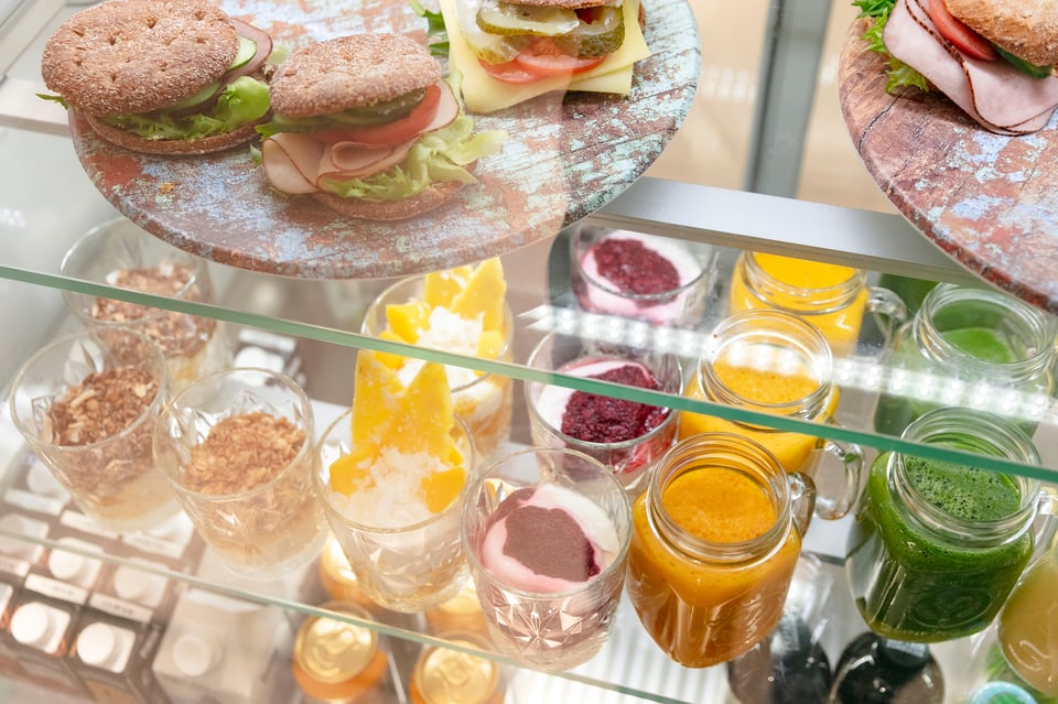 Kahvilan tarjoiluvitriini, jossa täytettyjä ruisleipiä, värikkäitä smoothieita, sekä erilaisia jogurttiannoksia granolalla, hillolla ja hedelmillä.