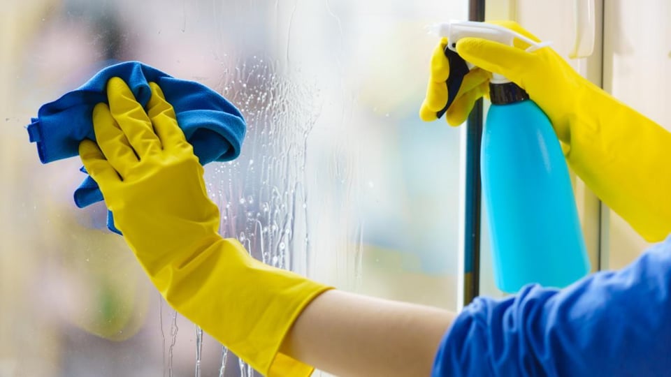 Kädet keltaisissa kumihanskoissa pesemässä ikkunaa suihkupullon ja siivousliinan kanssa.