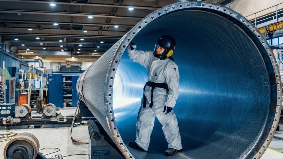 ISS:n siivooja suojahaalarissa puhdistamassa suurta metalliputkea tehdasympäristössä.