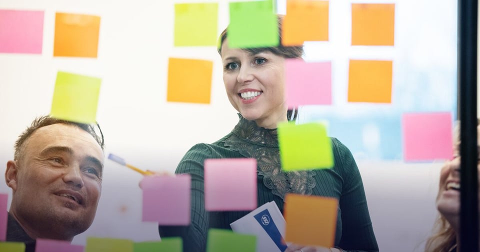 Nainen ja mies kokoustamassa lasin takana, joka peitetty värikkäillä post-it lapuilla. Nainen osoittaa post-it lappuja kynällään.