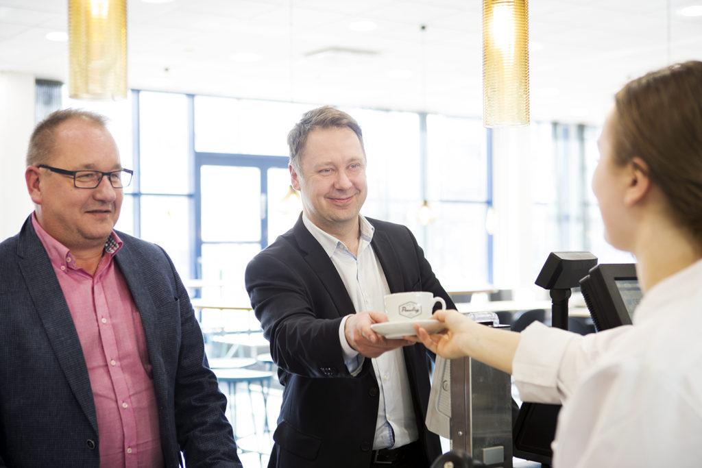 Varman Toni Pekonen ja Mika Kaasalainen kahvilan kassalla vastaanottamassa kahvikuppia.