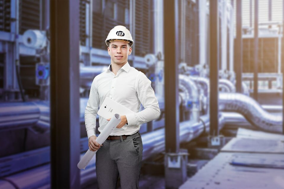 Mies ISS:n turvakypärässä ja kauluspaidassa kädessään paperiset piirrokset rullalla. Taustalla putkia.