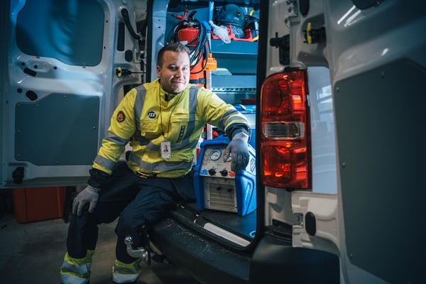 ISS:n kiinteistöhuoltaja istumassa huoltoauton takatilan edessä heijastintakissa ja työhousuissa. Auton takatilassa näkyvillä erilaisia työkaluja.