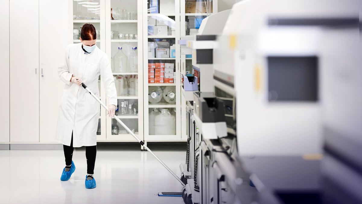 Siivooja valkoisessa työtakissa ja kasvomaskissa pesemässä lattioita Perkin Elmerin laboratoriossa.