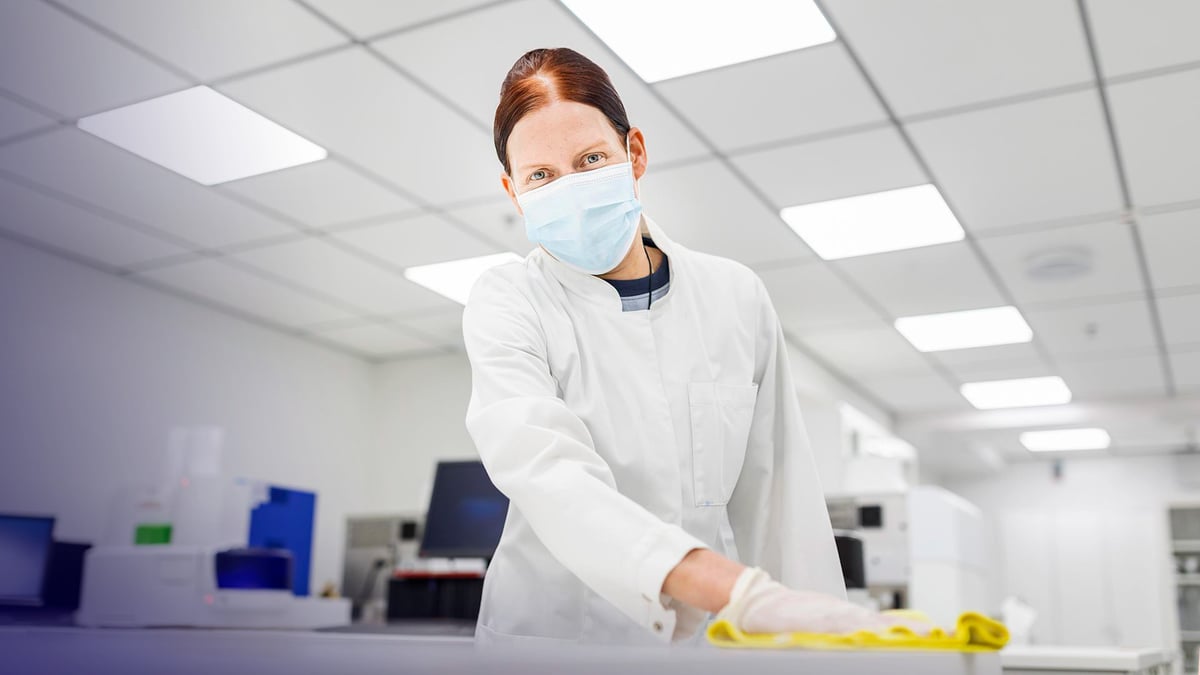 Siivooja valkoisessa työtakissa ja kasvomaskissa pyyhkimässä pöytää Perkin Elmerin laboratoriossa.